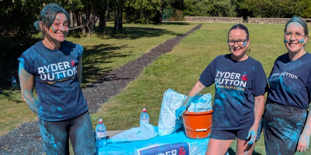 Three fundraisers in matching blue t-shirts with 'Ryder & Dutton' logo at an outdoor colour run event.