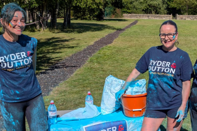 Three fundraisers in matching blue t-shirts with 'Ryder & Dutton' logo at an outdoor colour run event.