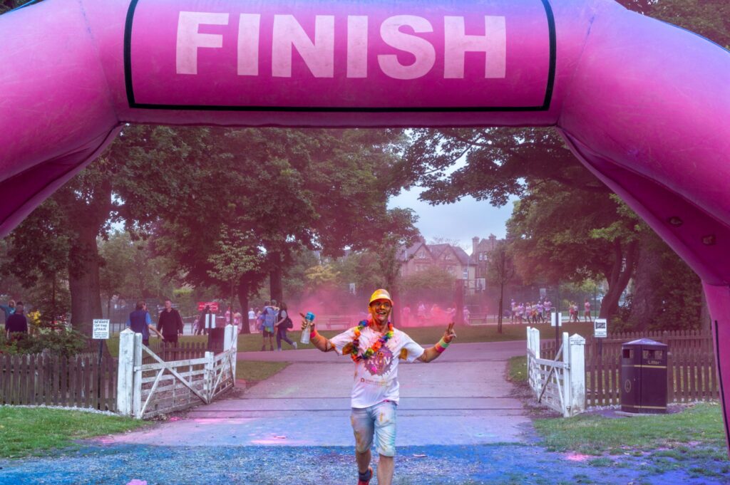 A man wearing a sun-visor and floral lei, is seen underneath a finish line, covered in colour powder after a rainbow run.
